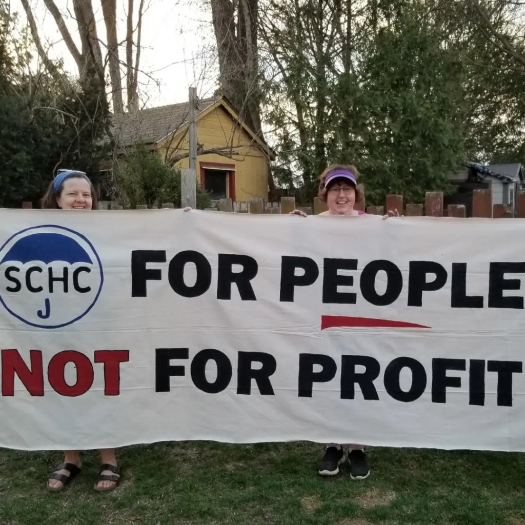 Two volunteers hold up a banner which reads: SCHC FOR PEOPLE NOT FOR PROFIT